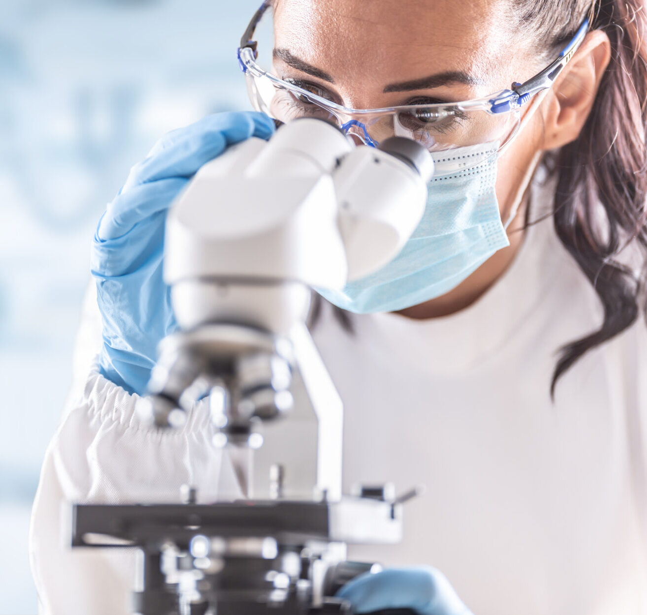 female in blue mask, white labcoat and wearing blue gloves, looking into a microscope