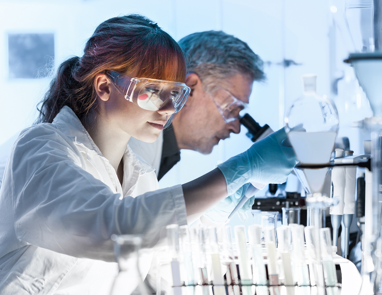 male with clear glasses looking into microscope alongside female with red hair in labcoat holding a sample vial
